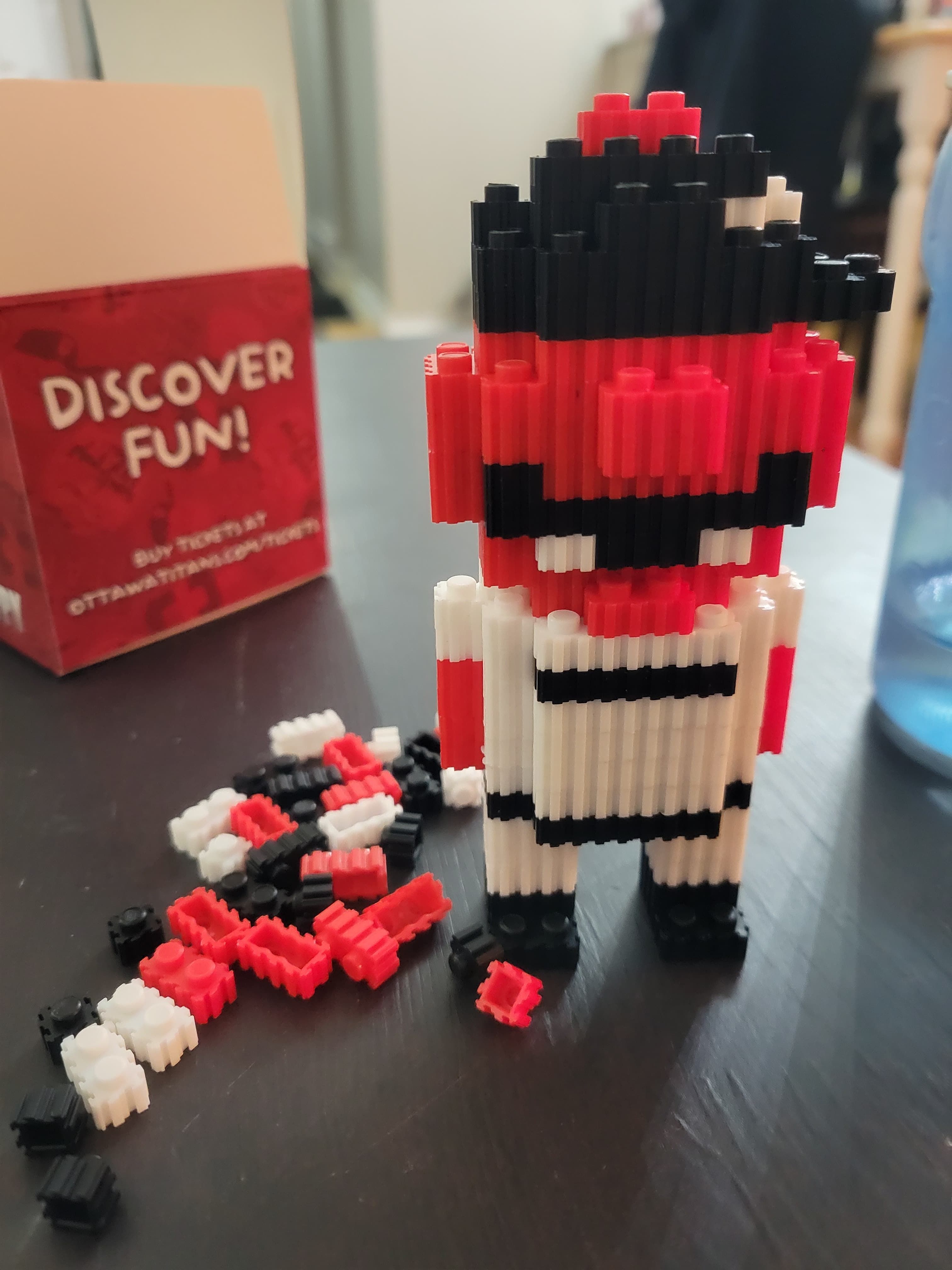 A block set version of Cappy, the Ottawa Titans mascot. The set is in three colours: white, black, and red. His head and arms are all red, with mouth and hat pieces in black. The rest is white. It is standing in front of a square box that says "Discover Fun!"