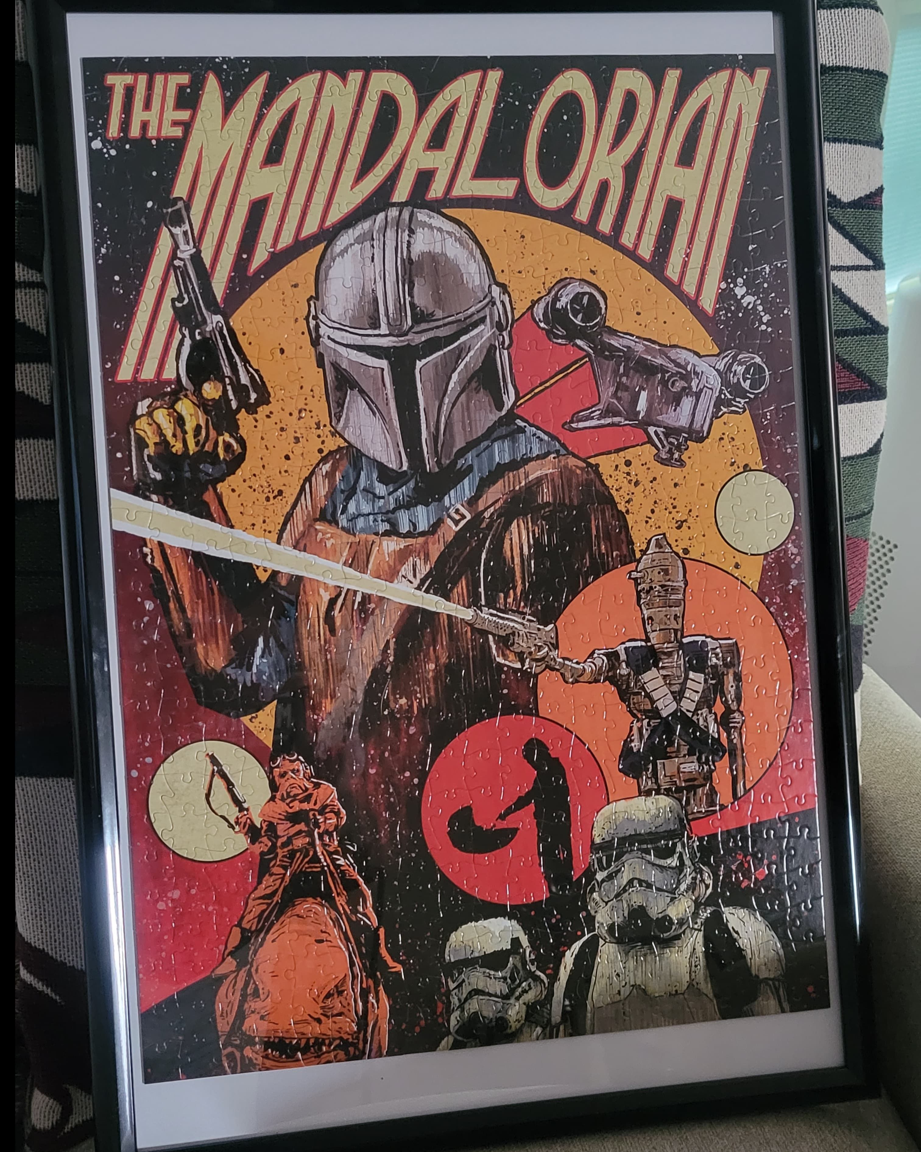 A completed, framed puzzle depicting stylized scenes from The Mandalorian TV show. The frame is thin with black borders.