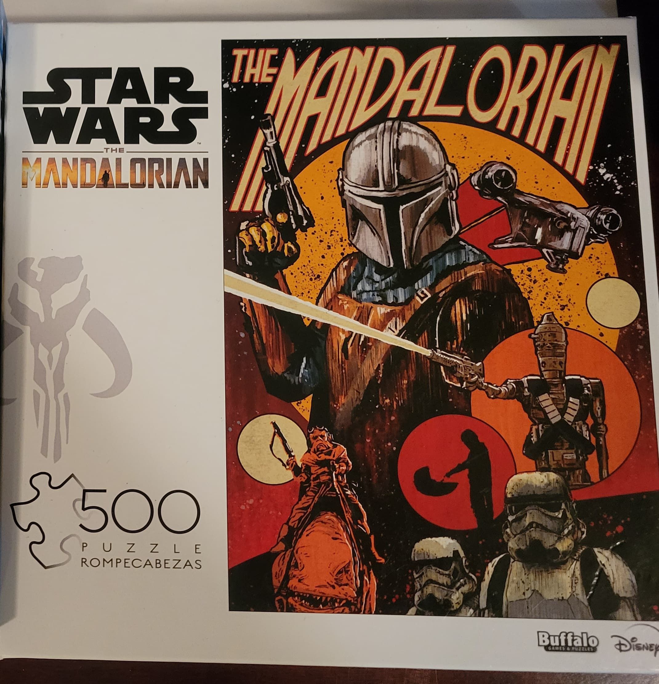 The box for a 500-piece puzzle by Buffalo games. The title is Star Wars The Mandalorian; the featured picture is the title character (The Mandalorian) in main focus with other side characters and scenes from the first season of the TV show.