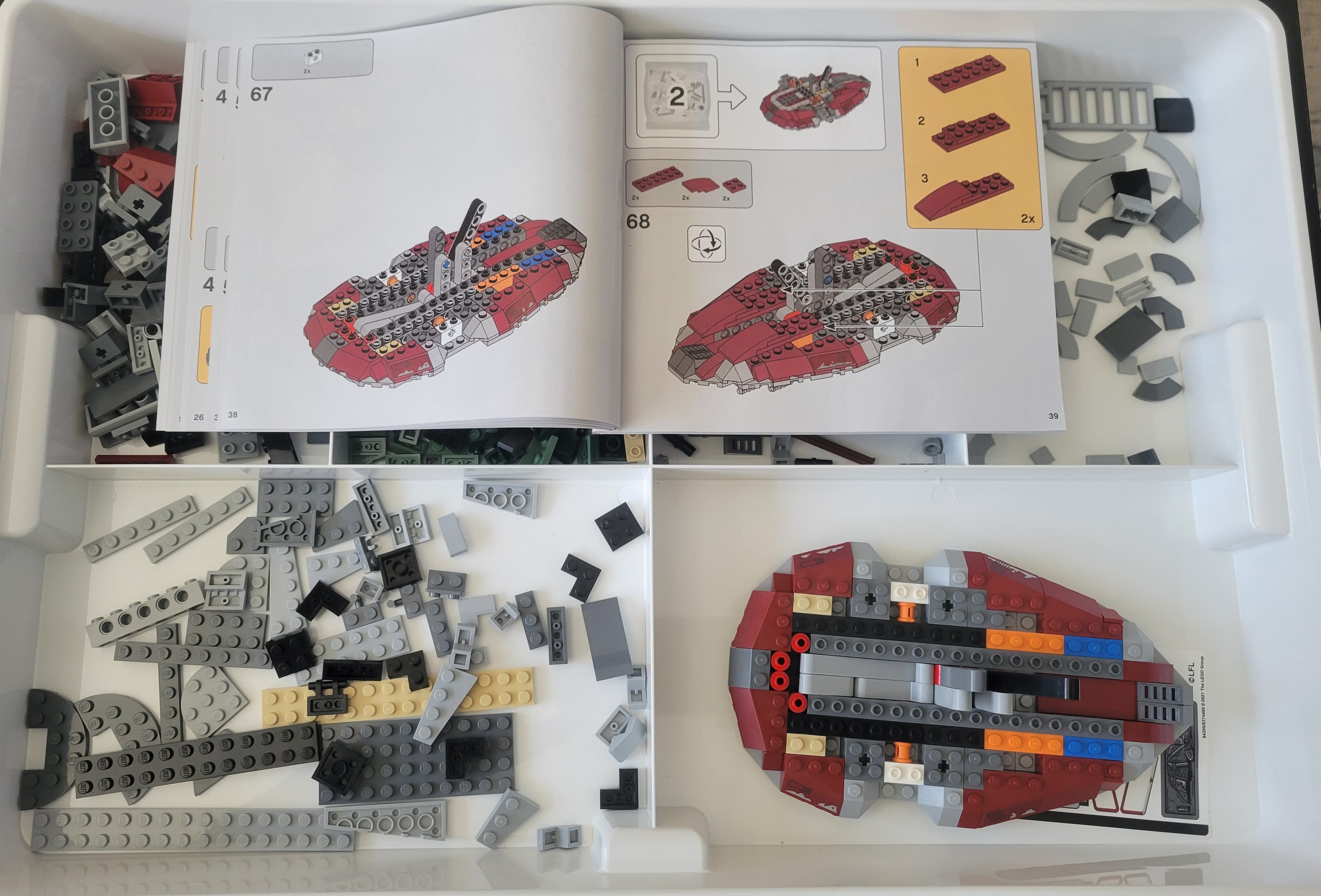 A LEGO instruction booklet sits atop a plastic tray containing various LEGO parts. The partially complete model of Boba Fett's Starship is in the bottom right of the tray.