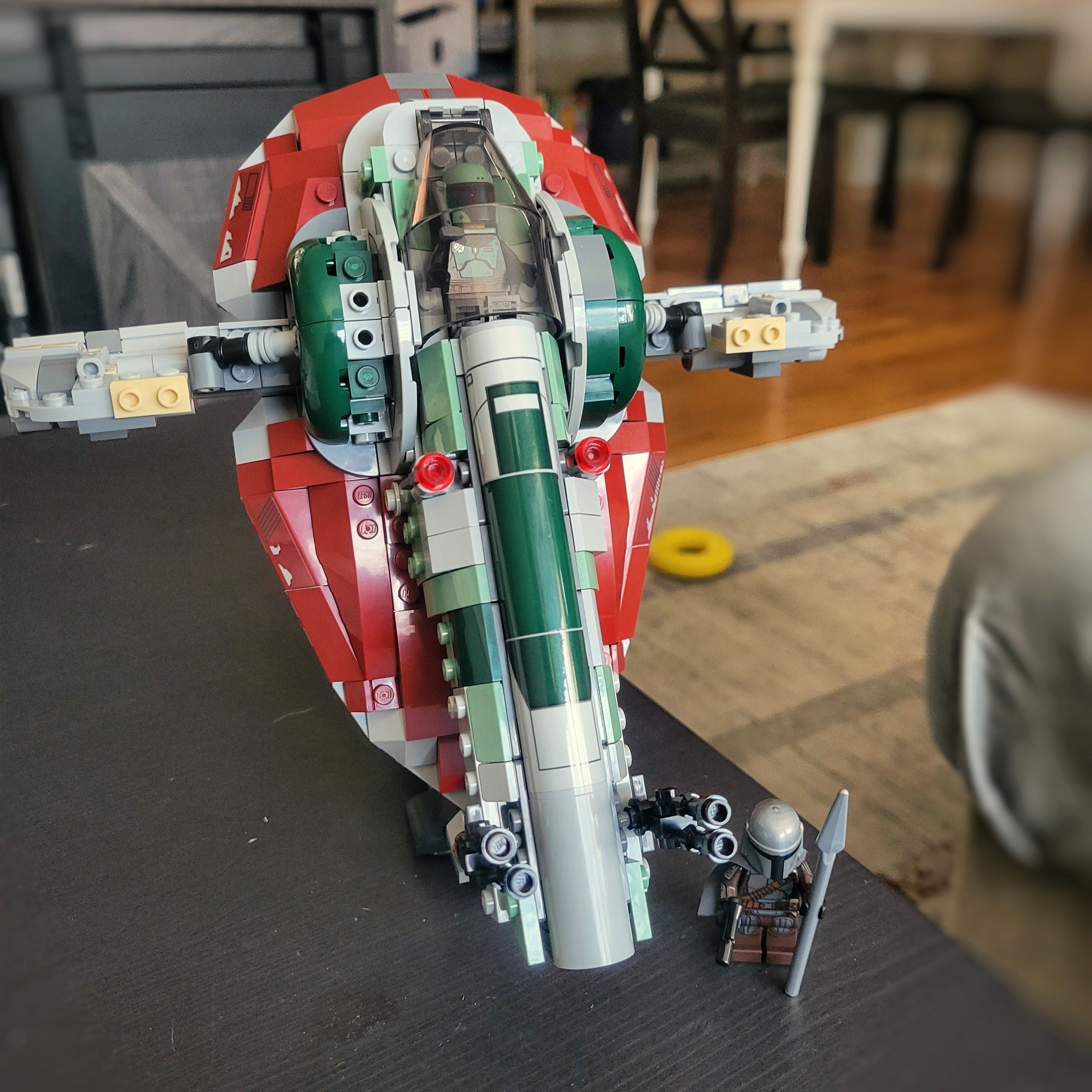 Boba Fett's Starship sits on a black tabletop. A Boba Fett minifig is inside the cockpit while a minifig of The Mandalorian, wearing predominantly gray armour, stands beside the ship.